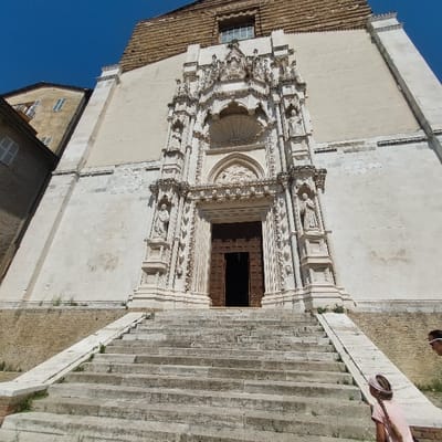 The image depicts the Cattedrale di San Ciriaco in Ancona, Italy, viewed from a low angle, emphasizing its grand entrance. The cathedral's facade is constructed of light-colored, weathered stone, with an upper section appearing to be of a different, textured material. A prominent, highly elaborate Gothic portal, sculpted from lighter white stone, forms the central focus. This portal features multiple carved arches, numerous niches housing statues of religious figures, and a detailed tympanum above the dark brown wooden double doors, which are partially open.

A wide, multi-tiered stone staircase, exhibiting signs of wear and minor vegetation growth in crevices, leads directly up to the portal. A stone ramp or low wall with a sloped top runs along the right side of the staircase. To the immediate left of the cathedral, a portion of an older, lighter-colored building with shuttered windows is visible. The sky above is clear and blue.

In the lower right foreground, two individuals are ascending the staircase. The foremost individual, a young female seen from behind, has light-colored hair styled in a braid or ponytail with a pink headband and is wearing a light pink top, looking upwards towards the cathedral entrance. Behind and slightly to her right, the top of another individual's head is partially visible, suggesting an adult possibly wearing sunglasses.