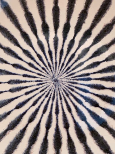 The image displays a close-up, abstract view of a patterned surface. The pattern consists of numerous dark blue, irregular lines radiating outwards from a central point, creating a sunburst or fan-like effect. The lines are not solid but appear to be made of textured, somewhat fuzzy strokes, giving them a slightly blurred or painterly quality. These lines alternate with sections of a light, off-white background. The background itself has a slightly uneven texture, with faint crackling visible, suggesting it might be ceramic or a similar material. The overall impression is a dynamic, geometric design with a handmade feel. There are no people, specific objects, or discernible scenes present. The focus is solely on the abstract pattern and texture of the surface. No information about time of day, weather, location cues, or emotions can be inferred from the image. There is no visible text.