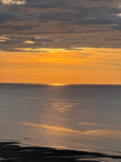 This is an aerial, high-angle, vertical shot of the ocean and sky during what appears to be sunset or sunrise. The horizon is clearly defined, separating the vast expanse of the ocean from the colorful sky. The ocean is a muted gray-blue, reflecting the warm tones of the sky in shimmering, golden streaks. There are dark, irregular shapes along the bottom edge of the frame, possibly a shoreline or rocky outcropping. In the distance, on the ocean's surface, small, dark specks are visible, suggesting the presence of boats. The sky is dominated by warm oranges and yellows near the horizon, transitioning to grayer, cloud-laden tones towards the top of the frame. The clouds are layered horizontally, with some streaks of light breaking through. The overall impression is one of a serene, natural landscape. The location is identified as Lý Sơn Special Administrative Region, Vietnam.