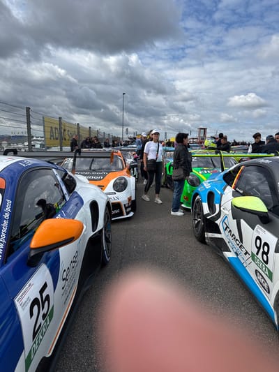 The image displays an outdoor scene at the Sachsenring circuit in Hohenstein-Ernstthal, Germany, under a partly cloudy sky. Multiple Porsche 911 GT3 Cup race cars are stationary on an asphalt surface, likely a pit lane or pre-grid area.
In the immediate foreground on the left, the blue, white, and orange side of a race car, numbered "25" and branded with "PORSCHE" and "DEKRA", is visible, featuring an orange side mirror. Directly to its right, an orange and white race car with "16" partially visible and "Gronau" branding is parked. Further right, a green and black race car, partially displaying "2" and "FIM" logos, is positioned. On the far right, the black, blue, and white side of a car with race number "98" and "TARGET Competition" and "DEKRA" branding is present, showing neon yellow accents.
Approximately fifteen to twenty individuals are standing on the asphalt among the race cars. A woman in a white t-shirt, white cap, and black pants is prominently featured in the mid-ground, looking right, with a lanyard visible. To her right stands another woman in a dark jacket and jeans, carrying a shoulder bag. Other individuals are scattered further in the background, some near the cars, others near the trackside barriers.
The background features metal catch fencing topped with chain-link fence, behind which a yellow banner with "ADAC" is partially visible. A small, red-and-white striped structure, possibly an observation post, is discernible in the far right background. The lower central portion of the image is obscured by a blurred human finger.