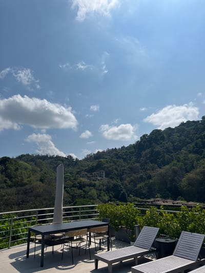 The image shows a patio overlooking a lush green hillside in Playa Herradura, Costa Rica. The patio features a black metal table with four chairs, a white umbrella, two lounge chairs, and a large potted plant. There are railings surrounding the patio, and the view from the patio shows a hillside covered in dense green trees and foliage. The sky is a bright blue with scattered white clouds. There are no people in the image. The scene is serene and peaceful, with the only sound likely being the chirping of birds and the rustling of leaves.