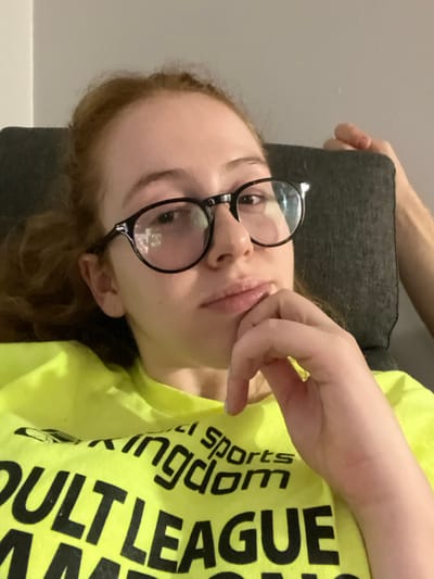 This image depicts a female individual in a close-up indoor shot, originating from Plainsboro Township, United States. The subject is a light-skinned female with reddish-brown hair pulled back, wearing round, dark-framed eyeglasses. Her expression is neutral, with lips slightly parted, and her left hand is positioned with fingers resting against her chin/jawline. She is wearing a neon yellow t-shirt with black text, partially visible, reading "KINGDOM", "Adult sports", and "ADULT LEAGUE". Behind her, a dark grey upholstered surface, likely a chair or sofa back, is visible, with a lighter grey wall or surface above it. A portion of another person's hand is visible on the upper right, gripping the top edge of the grey upholstered surface. The lighting suggests an indoor, ambient environment.