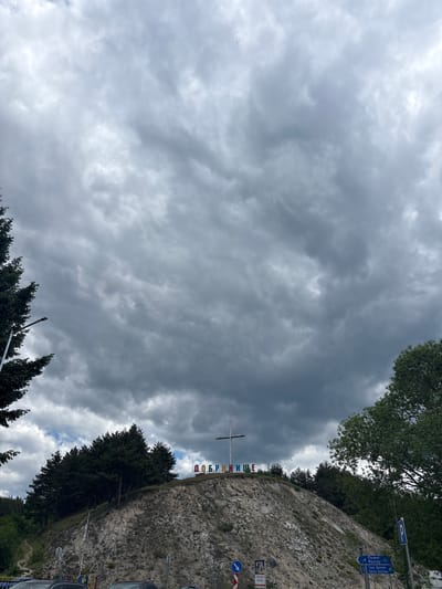 The image shows an upward view in Dobrinishte, Bulgaria, focusing on a cloudy sky dominating the frame. Below, a stone retaining wall is visible with green trees lining its edge. A large white cross with colorful letters spelling "ДОБРИНИЩЕ" vertically down the center stands prominently. A road sign showing the direction to Razlog, Toue Delchev, and Gotse Delchev is present along with a parking sign. A portion of a car is visible on the right edge.