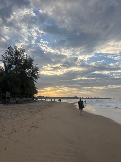 The image shows a beach scene at Arugam Bay, Sri Lanka. The sky is filled with dramatic clouds, with streaks of sunlight breaking through, suggesting either sunrise or sunset. The ocean is visible on the left, with gentle waves washing onto the shore. A sandy beach stretches out to the right, marked with footprints.
In the foreground, on the beach, a person is walking towards the water. They are wearing dark shorts and a dark t-shirt with white text, though it is difficult to read clearly. The person is holding a green object, possibly a surfboard or a paddleboard. Further down the beach, a line of people are visible walking along the shoreline, and some appear to be in the water. Lush green trees and palm fronds are seen on the left side of the image, adding to the tropical setting.
The overall mood is serene and peaceful, typical of a beach at this time of day. The lighting suggests a time of day with soft, warm light illuminating the clouds and the sand.