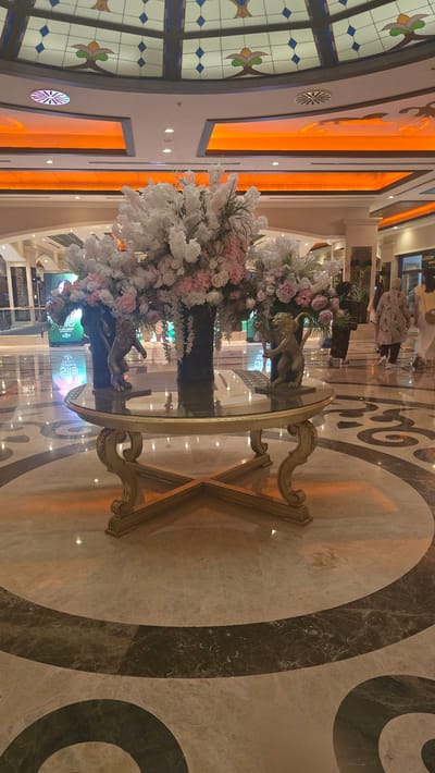 The image shows a grand interior space, likely a hotel lobby or shopping mall, in Kazafani, Northern Cyprus. In the foreground, a decorative round glass-top table with a gold ornate base stands on a marble floor with circular patterns. Atop the table are large floral arrangements featuring white and pink flowers. Two small golden lion statues are placed on the table, one on each side of the floral arrangements. The ceiling features a stained-glass dome and modern orange lighting panels. In the background, several people are walking, and shop signs are visible. The overall impression is one of luxury and opulence.