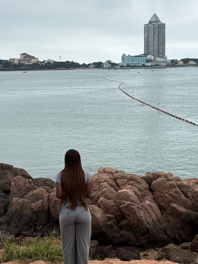 The image displays a coastal panorama within Shinan District, Qingdao, China, under an overcast, uniformly grey sky. In the immediate foreground, a person is seen from behind, standing on a rugged outcrop composed of reddish-brown rock formations and sparse green vegetation. The individual presents long, reddish-brown braided hair and wears a light grey short-sleeved t-shirt paired with grey loose-fitting pants. The person is oriented away from the camera, observing the scene across the water.
The midground features a calm, grey-blue bay. Two distinct lines of buoys are visible on the water; a closer, shorter line of dark buoys, and a longer, winding line of orange and white buoys marked with occasional small red flags extending towards the horizon. A small red paddle boat is discernible on the water near the distant shore.
The background reveals the urbanized coastline of Shinan District. Dominating the skyline is the tall, slender Qingdao International Financial Center, identifiable by its distinctive pointed, multi-faceted roof. Adjacent to it on the left stands a lower, light blue building complex. Further left, various other buildings, some featuring red-tiled roofs, are situated along the distant shore, with subtle indications of a beach area.