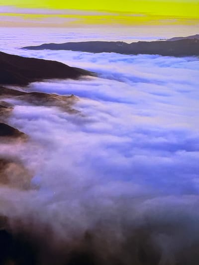 This image depicts a dramatic aerial view of a vast blanket of white and purple clouds, likely fog, filling a valley between dark, silhouetted mountain ranges. The clouds undulate and form distinct layers, with some areas appearing dense and opaque, while others are more translucent, allowing glimpses of the land beneath. A bright yellow-tinged horizon is visible above the clouds, suggesting either sunrise or sunset. The scene is devoid of any human presence or man-made structures, offering a purely natural landscape. The location is not identifiable as being within the city of Gorzow, Poland.
