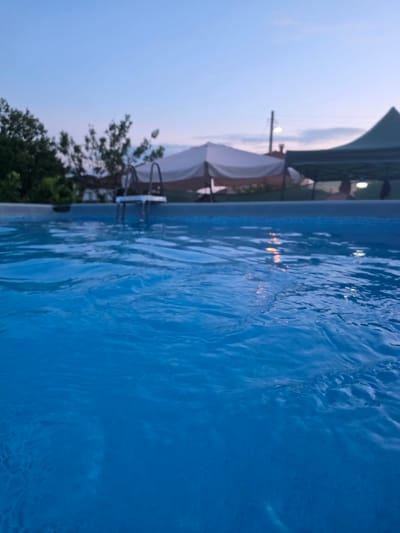 The image shows an outdoor swimming pool at dusk. The water is a vibrant blue and appears to be rippling. In the background, there are several tents or canopies, some of which are illuminated. Trees are visible to the left of the frame, and a ladder is positioned at the edge of the pool. The sky is a pale blue, indicating that the sun has recently set. There are no visible people in the immediate vicinity of the pool, but silhouettes of individuals can be seen within the tents in the background. The overall atmosphere suggests a recreational setting after dark. The location is specified as Tsaratsovo, Bulgaria.