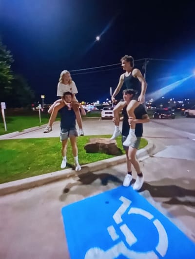 The image depicts a night scene in Baykaro, Uzbekistan, featuring four individuals in a paved outdoor area adjacent to a grassy verge. Two males, wearing t-shirts, shorts, and sneakers, are each carrying another person on their shoulders. On the left, a male in a dark t-shirt and light shorts supports a female with blonde hair, clad in a light-colored t-shirt and light shorts. The male on the ground holds a small object in his left hand. On the right, a second male in a dark tank top and dark shorts carries another male, who wears a dark t-shirt and dark shorts. All four individuals are illuminated by artificial light sources. In the immediate foreground, a blue rectangular marking with a white international symbol of access is painted on the pavement, indicating a designated accessible parking space. A concrete curb separates the paved area from a strip of green grass on the left. The background reveals multiple vehicle lights, streetlights, and illuminated commercial structures, consistent with an urban or developed suburban environment. Overhead power lines are visible against the dark sky.