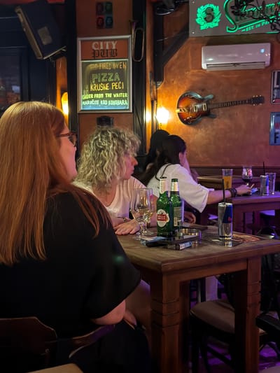 The image depicts a dimly lit interior of a pub, likely in Sarajevo, Bosnia and Herzegovina, given the context clues. In the foreground, a woman with long, reddish-brown hair, wearing glasses and a black top, is seated at a wooden table. To her right, another woman with curly blonde hair and a white top is also seated. On the table, there are several items, including two green bottles of Stella Artois beer, a Red Bull can, several glasses, an ashtray with cigarettes, and a menu. In the background, other patrons are visible, as well as decor that includes a neon sign with green lettering that reads "CITY PUB," and a framed sign displaying text related to pizza orders. An electric guitar is mounted on the wall above an air conditioning unit. The overall ambiance suggests a casual gathering at a local establishment.