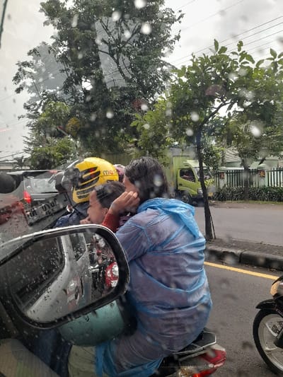 The scene depicts a street in Calukan RT 1 RW 5 Keboansikep, Indonesia, during rainfall, evidenced by numerous raindrops on the viewing surface and a passenger's attire. A motorcycle occupies the central foreground, carrying two individuals. The driver wears a yellow helmet with black stripes and partial text "max". The passenger, seated behind the driver, is clad in a translucent light blue poncho-style raincoat, with dark, wet hair; her left hand is raised near her ear.
In the lower-left, a rearview mirror reflects a sequence of vehicles, including a red scooter and a white car, indicating traffic behind the vantage point. Behind the central motorcycle, a dark car with a partially visible license plate is present. The far-right edge of the image shows the front wheel and engine component of a black motorcycle.
The background is characterized by an abundance of dense green trees under an overcast sky. A light green utility vehicle is discernible in the middle ground, partially obscured by foliage and a white fence structure. The road surface is asphalt, delineated by a yellow line and a raised curb. The overall environment suggests an urban or semi-urban setting experiencing active precipitation and vehicular movement.