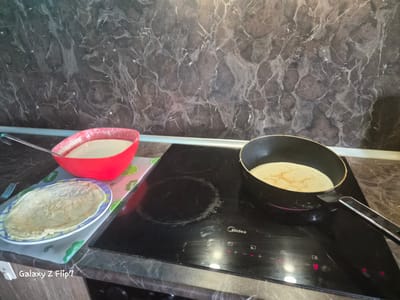The image captures a kitchen counter scene in Kalipetrovo, Bulgaria, depicting the process of making pancakes or crepes. The countertop features a dark, marble-patterned surface, which also extends as a backsplash along the wall behind it.

On the left side of the frame, a bright red mixing bowl is filled with light-colored pancake batter, with a silver ladle resting inside. Below the bowl, on a light-colored placemat decorated with green lime slices and the word "Lemon," rests a blue and white patterned plate holding a freshly cooked, light-brown pancake. A silver fork is visible to the left of the plate.

In the center and right, a black Midea induction stovetop is the focal point. A black frying pan sits on one of the hobs, containing a pancake currently cooking, showing light golden-brown marks. The stovetop's digital display shows the number '9', indicating an active and hot burner, along with other control symbols. The surface of the stovetop shows some smudges and residue from cooking.

The overall activity depicted is the preparation of pancakes. The scene suggests a continuous process, with batter ready, a pancake cooking, and another already finished and plated. The lighting appears natural, typical of an indoor setting during the day. A "Galaxy Z Flip7" watermark is present in the bottom left corner of the image.