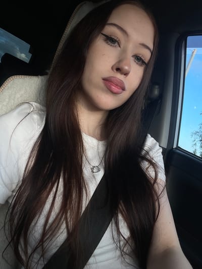 A young woman is captured in a close-up, selfie-style shot, taken from a slightly elevated angle. She has long, dark brown hair that cascades over her shoulders and chest. Her makeup is noticeable, with prominent eyeliner and full lips. She is wearing a white t-shirt, and a silver necklace with a pendant is visible. A black seatbelt is fastened across her body, suggesting she is in a vehicle.

The setting appears to be the interior of a car, with a glimpse of the sky and trees through an open sunroof. The light suggests it is daytime, possibly late afternoon, given the angle of the sun. The overall mood conveyed is relaxed and casual. No text is visible in the image. The location context provided is Gavrilovka, Russia.