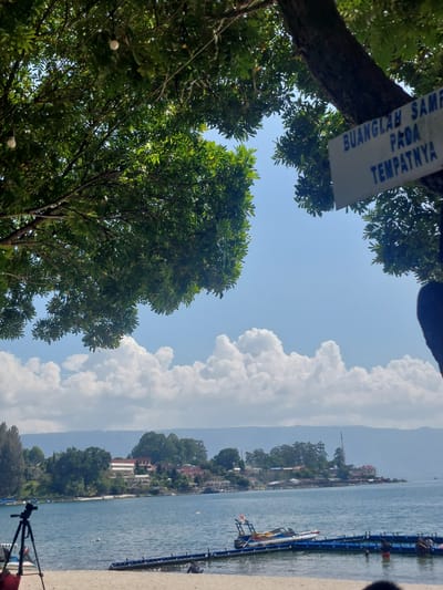 The image depicts a scenic view of a lakefront, likely in Parapat, Indonesia, on a clear day. In the foreground, a sandy beach is visible with a black tripod and a red bag. A blue and white floating dock extends into the water, where several people are seen swimming and interacting. A small boat is docked at the floating structure. The lake itself is calm and blue, with a distant shoreline featuring lush green trees and buildings, including what appears to be a resort or hotel. The sky is a bright blue, adorned with fluffy white clouds. Overhead, branches of a large tree frame the upper portion of the image, with two hanging light bulbs visible. A sign attached to the tree trunk reads "BUANGLAH SAMPA" which translates to "Dispose of trash". The overall atmosphere is tranquil and evokes a sense of leisure and natural beauty.