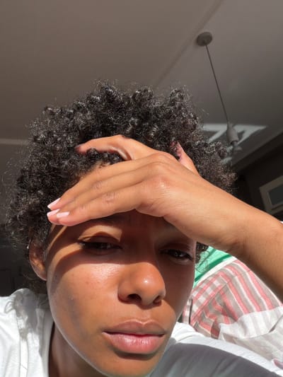 In a close-up, indoor shot, a person with dark, curly hair rests their forehead on their right hand, which is positioned across their brow. The individual's left eye and partial view of their nose and lips are visible, illuminated by a strong, directional light source casting a shadow across their face. The background is indistinct, with hints of striped fabric and a ceiling fixture with a shadow pattern suggesting a slanted roof or window. The scene appears to be within a residential setting.