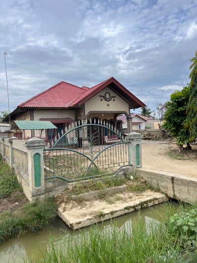 The image depicts a residential property in Bundar, Indonesia. The foreground features a narrow, muddy canal with overgrown green grass along its banks. A concrete pathway leads from the canal towards a house, with steps made of concrete. A decorative metal gate, painted green, is a prominent feature of the property's fence, which is constructed from concrete pillars and horizontal bars, also painted green. The house itself has a prominent red tiled roof and a beige-colored facade. A smaller green awning is visible on the left side of the house, and a larger entrance with decorative columns is on the right. The sky is overcast with clouds. In the background, other houses and trees are visible, suggesting a semi-rural or suburban setting.