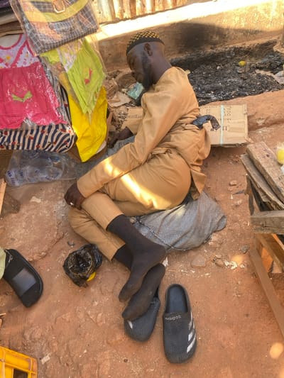 An adult male is observed sleeping outdoors in Sabon Gari South, Nigeria. He is seated on a grey patterned fabric on reddish-brown earth, leaning against an unseen support. The male wears a light brown long-sleeved tunic and matching trousers, dark socks, and a yellow and black checkered kufi-style cap. His eyes are closed, and his head is tilted to the right. A dark-colored object is tucked into the waistband of his trousers at the lower back. A silver-toned ring is visible on his right hand. Two dark grey textured slides, one bearing the Adidas logo, rest on the ground in front of him, along with a partially visible black slip-on shoe to the left. A small black plastic bag containing an unknown yellow object is also on the ground near his feet.
To the left of the male, a vertical stack of merchandise, including colorful packaged clothing and clear plastic bottles of "ABU-DANNA" brand water, is visible. To his right and rear, a cardboard box is present, partially obscuring an area of dark, possibly burnt, material on the ground. Stacked wooden planks are visible further to the right. The background features corrugated metal sheeting. The scene is illuminated by natural sunlight, casting distinct shadows.