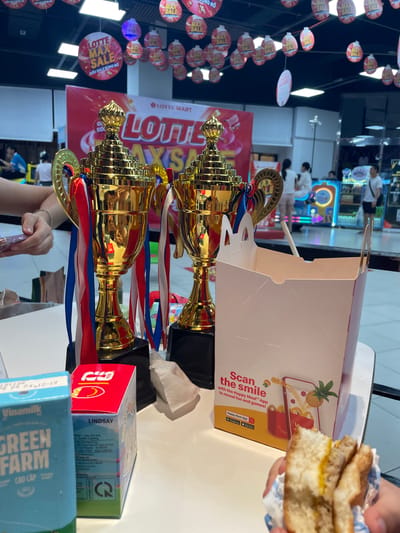 In Thuận An, Vietnam, two golden trophies with red, white, and blue ribbons stand on a white table, positioned in front of a large red banner that reads "LOTTE MART LOTTE MAX SALE." To the right of the trophies is a white cardboard box with "Scan the smile with the Happy Meal App" printed on it. In the foreground, a person is holding a partially eaten sandwich. Behind the table, several people are visible in the background, and the ceiling of a store, possibly a supermarket, is illuminated with lights. Red circular decorations with "LOTTE MAX SALE" printed on them hang from the ceiling.