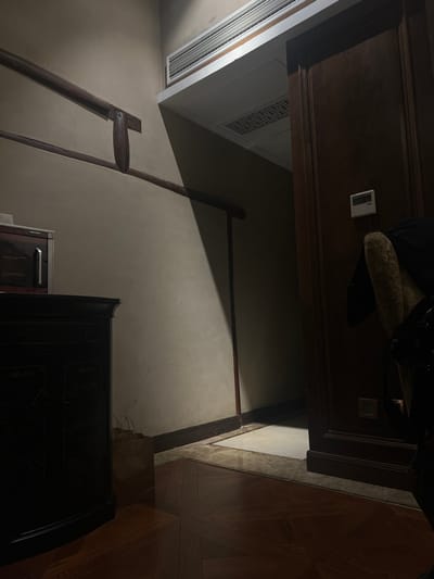 This image depicts an interior space in Tongxiang, China, characterized by dim, directional illumination. On the left, a dark, ornate wooden cabinet with carved panels supports a compact metallic appliance with a reflective front. A light-toned wall, extending from left to center, features two parallel dark wooden railings mounted horizontally. A strong diagonal beam of light originates from an unseen source, brightly illuminating a section of this wall and the adjacent floor, while casting a prominent shadow across the upper right wall and the area beyond. The floor transitions from a dark wooden parquet pattern in the foreground to a lighter, possibly tiled or marble, surface leading into an open dark wooden door on the right. A white digital thermostat or control panel is affixed to the wall beside the door. Above the doorway, a metallic air conditioning vent is visible near the ceiling. Further into the shadowed space beyond the door, a ceiling panel with a decorative grid pattern is discernible. No individuals are present in the scene. A beige, textured object, partially obscured by deep shadow, is visible on the far right edge of the frame.