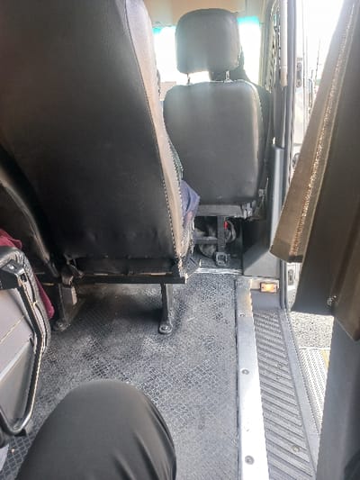 Inside an industrial vehicle, the interior features black leather seats with adjustable headrests. A textured gray floor extends along the right side, bordered by a silver metal strip. A metal bar runs above the seats. A small, illuminated orange light fixture is visible along the gray textured wall. A dark-colored bag sits beneath the seats.