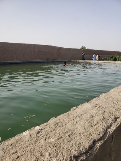 A daytime shot of a canal or large concrete trough. Three people stand along the left bank, while one swims in the canal. The banks are concrete or mud brick. The water has a green tint and some debris is floating in it. The location appears to be Arghandab, Afghanistan.