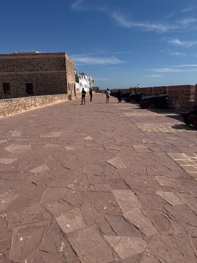 The image shows a sun-drenched scene on a historic fort's ramparts, likely in Essaouira, Morocco, given the location context. The ground is paved with large, irregular reddish-brown flagstones, reflecting the bright sunlight. Along the left side of the rampart, a stone wall with visible stonework forms the fort's structure, with a few dark-colored windows. Several cannons are mounted on the wall, pointing outwards, suggesting a defensive position with a view over a body of water, possibly the ocean.
The sky is a clear, bright blue with sparse, wispy white clouds, indicating a fair weather day. Several seagulls are visible in the sky, one soaring near the cannons.
On the rampart, a few people are walking and standing, appearing to be tourists exploring the fort. They are casually dressed, enjoying the warm weather and the historical surroundings. The figures are small in the frame, emphasizing the grandeur of the fort and its expansive setting. In the background, white buildings with traditional architecture can be seen, possibly part of the city adjacent to the fort. The overall mood is peaceful and observational, capturing a moment of exploration and enjoyment of a historical site on a pleasant day. No visible text is present in the image.