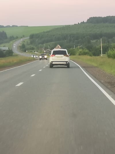 The image depicts a road in Забегалово, Russia, with a white SUV in the foreground, traveling away from the camera. Several other vehicles are visible on the winding road ahead, including a dark-colored sedan with its headlights on. The road is asphalt, with white lane markings. The surrounding environment consists of rolling green hills covered in trees and fields, with scattered houses and structures visible on the left side of the road. The sky is a pale, hazy color, suggesting it might be dusk or dawn. There are no people clearly visible in the image.