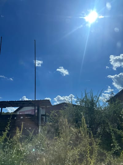 In Konstantinovo, Bulgaria, a bright sun illuminates a clear blue sky dotted with fluffy white clouds. Two tall, slender poles stand in the foreground, with one extending higher than the other. Beneath the sky, a rustic wooden structure with a corrugated roof is visible, partially obscured by lush green foliage. A sign with Cyrillic lettering, "Денислав," is affixed to the front of the building. The scene is bathed in sunlight, creating a warm and vibrant atmosphere.