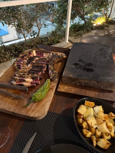 An outdoor dining scene in Nardò, Italy, features a prepared meal on a dark wooden table during evening hours. Dominating the center-left is a rectangular wooden cutting board holding a deconstructed T-bone steak, with the meat sliced and fanned out beside its bone, displaying a medium-rare interior. A single green chili pepper and a pair of silver metal tongs are placed on the board. To the right, a thick, dark grey hot stone for cooking, showing some residue, rests on a smaller wooden base. In the lower right, a black oval bowl contains roasted, golden-brown potato cubes. The table is set with a black textured placemat, a partially visible clear glass on the left, and a serrated knife blade. The background reveals lush green foliage illuminated by warm artificial lights, alongside white vertical and horizontal structural elements consistent with a patio or balcony railing. No individuals are visible in the frame.