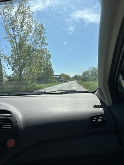 The image, taken from inside a vehicle looking out the front windshield, captures a bright, sunny day on a rural road in Voysil, Bulgaria. The perspective is from the driver's or passenger's seat, angled slightly to the right, showing the road stretching into the distance.

**Subjects:** The primary subjects outside the vehicle are a paved road, bordered by vibrant green trees and lush vegetation. A large deciduous tree with abundant green leaves dominates the right foreground and midground. In the distance, several vehicles, appearing to be white cars or vans, are visible on the road. Faint outlines of buildings or houses can be discerned beyond the trees. The sky is a clear, brilliant blue with sparse, wispy white clouds. Inside the vehicle, parts of the dark-colored dashboard are visible, featuring a textured surface and air vents. A circular red sticker is affixed to the right side of the dashboard. The windshield itself shows small specks, possibly dust or reflections.

**Activity or event taking place:** The scene suggests the vehicle is either in motion or has recently stopped along the road, offering a view of the passing countryside. It conveys the experience of a journey through a scenic, open area.

**Notable details:** The time of day is clearly daylight, indicated by the bright illumination and the blue sky. The strong shadows on the dashboard and the vivid green foliage suggest a sunny spring or summer day. The setting appears to be a tranquil, rural or semi-rural landscape.

**Visible text:** No discernible text is visible on any objects, vehicles, or signs within the image.