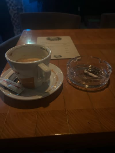 A table setting features an ashtray with a cigarette butt, a menu, and a cup on a saucer. The cup appears to contain a beverage. A small package of creamer and a spoon rest on the saucer. The table has a brown, wooden surface with visible seams. Lighting is dim, with darker areas to the right of the frame.