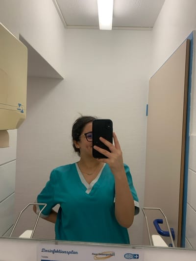 A female individual is observed in a mirror selfie taken within what appears to be a lavatory or hygiene area of a medical or care facility. She is positioned centrally, facing the mirror, and holds a black smartphone with her right hand, obscuring the lower half of her face. Her left arm is slightly bent with her hand on her hip. She has dark hair pulled back and wears eyeglasses. Her attire consists of a teal-colored medical scrub top featuring a white V-neck collar and white trim on the sleeves. A thin, delicate necklace is visible around her neck.

The environment is brightly lit by a rectangular fluorescent light fixture mounted on a white ceiling. The walls are white with a subtle textured pattern. To the reflected left, a beige-colored wall-mounted dispenser, likely for paper towels or hand sanitizer, is visible. To the reflected right, a light-colored door or partition with a prominent blue trim and a blue U-shaped grab bar is present. Below the mirror, a white surface serves as a counter. A sign is visible at the bottom edge of the mirror, featuring the text "Desinfektionsplan" (Disinfection Plan) and "Toilette" (Toilet). The sign also displays the name "Haus Geborgenheit" and a "WIBU+" logo.

Based on the visible signage, the location is identified as Haus Geborgenheit, a care facility situated in Neuffen, Germany.