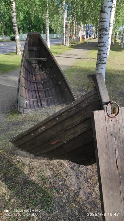Two old wooden boats are positioned outdoors in a park-like setting in Iisalmi, Finland. One boat is in the foreground, appearing to be partly submerged in the ground and resting on a wooden plank. It is dark brown and weathered, showing signs of age and wear. The other boat is in the background, also wooden and dark brown, positioned at an angle on a path. The boats are surrounded by green grass and a path, with birch trees and other foliage in the background. In the far background, there are playground-like structures with colorful ropes and poles. The sky is overcast. The image includes a timestamp in the lower right corner indicating July 8, 2026, 18:43.