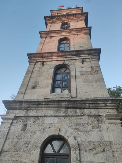 This is a low-angle, close-up, eye-level, upward shot of a stone clock tower under a clear blue sky. The tower is tall and appears to be constructed from large, light-colored stone blocks. It has a tiered structure with recessed sections and decorative stone moldings. Several arched windows with dark wooden frames are visible at different levels of the tower. Some of these windows have panes that reflect the sky and trees. A small red flag flies at the very top of the tower, above a railing. The sunlight suggests it is daytime, likely late afternoon or early morning given the direction of the light and the warm tones of the stone. The scene is set outdoors, and the presence of trees in the background indicates a natural or park-like environment. The overall impression is one of historical architecture bathed in natural light. There is no visible text in the image. The location is Osmangazi, Turkey.