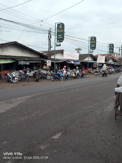 The image shows a street scene in Kecamatan Anjatan, Indonesia, with a row of motorcycles parked along the side of the road. The street is wet and appears to be paved with asphalt. There are several buildings visible on the left and right side of the street, with signs advertising a shop called "Setya". Several people are visible in the background, including a person standing behind a table and another person standing near a motorcycle. The time of day is mid-morning, with overcast skies. The motorcycles are parked in a single line on the right side of the street, facing the camera. The street is relatively empty, with only a few people visible in the distance.