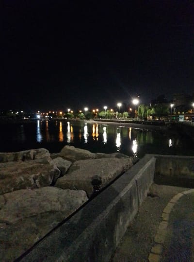 The image shows a nighttime view of a waterfront promenade in Kadıköy, Turkey. In the foreground, a rough stone wall lines the edge of a pathway. Beyond the wall, a body of dark water reflects the lights of streetlamps and buildings visible along the distant shoreline. Trees and structures are silhouetted against the night sky, with several bright streetlights illuminating the area. The water's surface is calm, with distinct reflections of the lights creating streaks of illumination. The overall scene depicts a tranquil, urban waterfront at night.