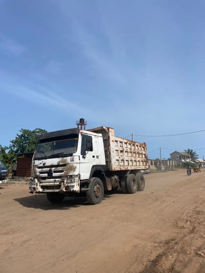 A white, heavily rusted dump truck is parked on a dirt road in what appears to be Ikoga, Nigeria. The truck is facing the right side of the frame. The truck bed is visibly corroded. The truck's front is heavily damaged and rusty. To the right and behind the truck are buildings and a person on a motorcycle. A tree and corrugated metal sheeting are on the left of the truck. The sky is clear and blue.