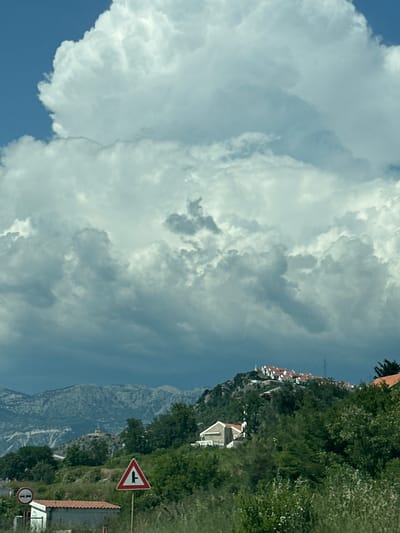 This image captures a scene in Blizikuće, Montenegro, under a dramatic sky filled with large, billowing white and gray clouds. The clouds occupy the upper two-thirds of the frame, with the lower portion featuring a landscape. In the mid-ground, a hillside is covered with lush green trees and vegetation. Several houses with red-tiled roofs are clustered on the crest of this hill, forming a small settlement. To the left of the hill, a building with a red roof is visible. In the foreground, a traffic sign with a red triangle and a black intersection symbol stands on a pole. Another circular traffic sign with a red border and a horizontal bar is partially visible to the far left. The background displays a range of rugged, grey mountains. The overall impression is one of a coastal mountainous region with a developing settlement, under an imposing and overcast sky.