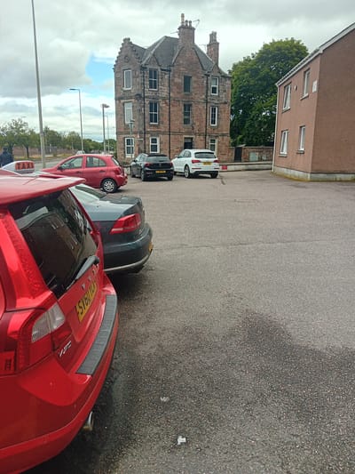 A paved parking lot is shown in the city of Inverness, United Kingdom, with several cars parked and a multi-story brick building in the background. The building is of traditional stone construction with several windows and a distinctive pitched roof. To the right of the brick building is a modern, pink-colored building with white-trimmed windows. In the foreground, a red Volvo wagon is parked closest to the viewer. Behind it is a dark grey sedan. Further back, a white Audi SUV is parked, followed by a red hatchback with a "TAXI" sign on its roof. A person in a dark jacket is walking away from the camera near the taxi. The asphalt of the parking lot appears wet in places, suggesting recent rain. The sky is overcast with patches of blue.