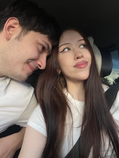 The image shows a close-up, selfie-style shot of a young couple lying down, possibly in a car. The woman is on top, her head resting on the man's shoulder. She has long, dark brown hair and is looking upwards with a slightly dreamy expression, her lips parted. She is wearing a white t-shirt and has dramatic winged eyeliner. The man is looking up at her, smiling slightly. He has dark hair and stubble. Both are wearing white t-shirts. The background is dark and out of focus, but suggests an interior space, perhaps a car, with a hint of sky and trees visible through a window. The lighting suggests it is daytime. No text is visible. The image conveys a sense of intimacy and affection.