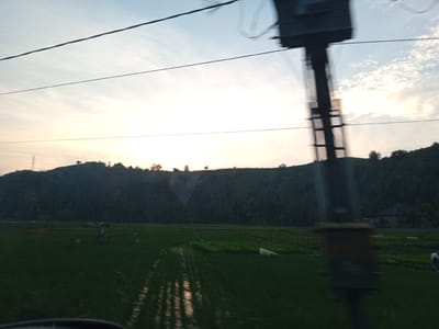 The image captures a rural Indonesian landscape at dusk, likely in Uleejalan, Indonesia. The foreground is dominated by a flooded rice paddy, reflecting the soft light of the setting sun and the sky. The water is disturbed, suggesting movement, possibly from wind or being traversed. Beyond the paddy, a dense, dark green hillside rises, covered in trees, and culminating in a ridge against the horizon. The sky above is a gradient of soft blues and pale oranges, with wispy clouds scattered across it. In the mid-ground, a rudimentary wooden structure, possibly a scarecrow or a small shelter, stands in the paddy. Further in the distance, a cluster of buildings, including a house with a dark roof, is nestled amongst the trees on the hillside. A tall, dark, blurry structure, possibly a utility pole or part of a vehicle, obstructs the right side of the frame, and thin, dark lines, likely power lines, stretch across the upper portion of the image. The overall impression is one of natural beauty and agricultural life in the twilight.