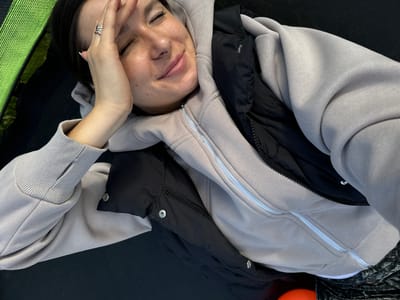 A woman lies on a black trampoline in an outdoor setting, possibly in Vysokinichi, Russia. She is wearing a light gray hooded sweatshirt over a black padded vest. The woman's eyes are closed, and she is smiling with her right hand on her forehead, which has a silver ring on one of the fingers. Green trampoline springs are visible in the upper left.