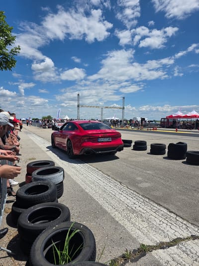 On a sunny day in Akatsievo, Bulgaria, a vibrant red Audi RS7 is captured in motion on a race track. The car, with license plate EH 9777 MB, is positioned on the right side of the track, facing away from the viewer and slightly to the left. Its sleek design and sporty stance suggest it is either accelerating or preparing for a race.

Surrounding the car, several stacks of tires are scattered on the asphalt, indicating a potential drag racing event or a practice session. The track itself is wide and appears to be made of asphalt, with white lines marking the lanes. In the background, a crowd of spectators lines the track, some standing and others seated behind a metal barrier, suggesting an organized event. Tents and temporary structures are visible further down the track, possibly for vendors or event organization.

The sky is a brilliant blue, dotted with fluffy white clouds, creating a picturesque backdrop. A few trees are visible on the far left of the frame, adding a touch of nature to the otherwise man-made environment of the race track. The overall scene depicts a dynamic outdoor event, likely a car race, set against a clear and sunny sky.