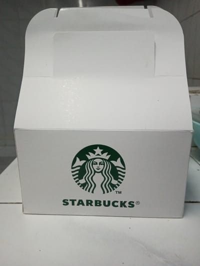 A white, glossy cardboard box, branded with the green Starbucks Siren logo and "STARBUCKS®" text, is positioned centrally on a light-colored, textured surface. The box's top flap is partially open, with its rounded side panels and central tab visible, indicating an unsealed state. Minor dark specks are visible on the box surface and the supporting surface. The background is out of focus, revealing a lighter wall or tiled surface, with a faint blue-green object visible on the right. No individuals are present. This scene is located in the city of B. Nam Ngao, Laos.