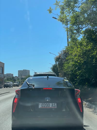 A black car is in the foreground, its rear end facing the viewer. The license plate is visible and reads "0200 AS GE". The car is in traffic, with other vehicles ahead and behind it. Tall green trees with lush foliage are on the left side of the image, casting shadows on the road. The sky is a clear, bright blue, suggesting a sunny day. In the distance, several multi-story buildings are visible, hinting at an urban or suburban setting. The perspective of the photo, taken from inside another vehicle, implies the viewer is also in traffic. The scene appears to be set in Aktau, Kazakhstan, during daylight hours with pleasant weather. There are no people clearly visible. The overall mood is neutral, characteristic of everyday traffic.
