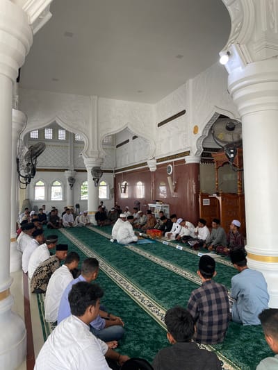 This media file presents an interior view of Mesjid Reubee in Indonesia, filled with men engaged in a religious gathering. The image is oriented vertically.
The scene features numerous men, predominantly young to middle-aged, seated on a green carpet with decorative patterns. Many are wearing traditional Muslim attire, including "koko" shirts and "peci" or "songkok" hats. Several individuals have books, likely religious texts or Qurans, open on small stands or on the carpet in front of them, suggesting they are participating in a study session, recitation, or listening to a sermon. Their postures indicate attentiveness and reverence.
The mosque's interior is brightly lit by both artificial overhead bulbs and natural light entering through arched windows. The architecture is prominent, featuring large white columns, ornate arches, and intricate white plasterwork with floral and geometric designs on the walls and ceilings, accented with gold. Several electric fans are visible, providing air circulation. In the background, a wooden minbar (pulpit) can be seen, along with a wall clock. A sign near the minbar displays the text "KHUTBAH MAKSIMAL 20 MENIT," which translates to "Sermon Maximum 20 Minutes." The overall setting conveys a peaceful and devout atmosphere during what appears to be a daytime religious event.
