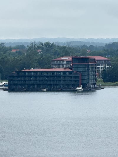 The media file displays a large, dark-colored, multi-story building situated along the edge of a body of water. The building has a prominent red roof and appears to be a hotel or similar establishment. The architecture is modern, with numerous windows. To the left of the building, there is a lush green hillside covered in trees, with some smaller buildings with red roofs visible amongst the foliage. In the distance, hills fade into a hazy, overcast sky, suggesting a possibly humid or misty atmosphere. The water in the foreground is calm and appears to be a lake or a wide river. Several small white structures, possibly boats or docks, are visible along the water's edge near the building. The overall scene depicts a waterfront property nestled within a natural landscape, under a muted sky. The orientation of the image is rotated 90 degrees counter-clockwise. There is no visible text in the media file. The time of day appears to be overcast, and the weather is likely humid or misty, given the haze in the background. The location is Mikołajki, Poland, a town known for its lake district.
