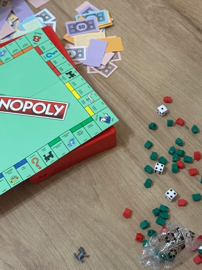 An overhead shot captures various components of a Monopoly board game scattered across a light brown wooden surface, likely a table or floor. The scene suggests a game either recently played, in progress, or about to begin.

In the center-left, a vibrant green Monopoly board with a red border is partially visible, featuring the prominent white and red "MONOPOLY" logo. Visible property spaces and elements on the board include "LUXURY TAX", "PARK PLACE", "CHANCE", "SHORT LINE", "COMMUNITY CHEST", "PENNSYLVANIA AVENUE", "NORTH CAROLINA AVENUE", "PACIFIC AVENUE", "GO TO JAIL", "MARVIN GARDENS", "ATLANTIC AVENUE", "VENINOR WORKS", "FREE PARKING", and a "BRAND" square. Various monetary values like "M200", "M300", "M320", "M350", "M280" are also present.

A pile of colorful Monopoly money is strewn across the bottom-left corner, featuring denominations such as "50" (purple) and "1" (light blue), alongside other light yellow, orange, and pink bills.

Scattered across the top and right side of the wooden surface are numerous red hotel pieces and green house pieces. Two white dice with black pips are visible among them; one displays a '5' and a '2', while the other shows a '6' and a '3'. A crumpled clear plastic bag containing more red hotels and green houses sits in the top-right. Text on the bag, possibly mirrored or partial, includes "EUOTE", "otni.9loy3915worl", "Cove", "Drob-f", "2016", "CVHYDV", and "nav". A silver-colored metal game token, shaped like a Scottie dog, is isolated in the far right of the frame. The lighting appears to be ambient indoor light.