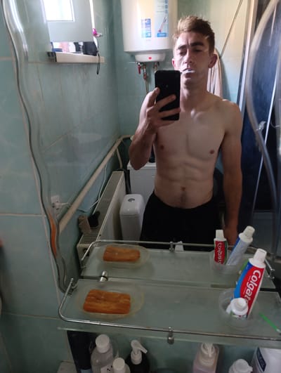 In a bathroom, a shirtless young man with short brown hair and visible abdominal muscles is reflected in a mirror. He is holding a black smartphone in his left hand and a toothbrush in his mouth. He is wearing black shorts. The bathroom features light blue tiled walls, a white water heater, a white toilet, a radiator, and a mirror with two glass shelves. On the shelves, there are two pieces of orange soap, tubes of Colgate toothpaste, and other toiletries. The lighting suggests it is daytime. The location is indicated as Fastiv, Ukraine.