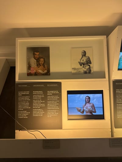This is a photograph of an exhibit in the Bernisches Historisches Museum in Bern, Switzerland. The exhibit features images and video related to Ursula Andress and her role as Honey Ryder in the James Bond film "Dr. No." In the background, there are two framed images: one is a movie poster featuring Sean Connery and Ursula Andress, and the other is a black and white photograph of Andress. In the foreground, a video is playing on a screen, showing Andress as Honey Ryder emerging from the water. Text panels in German and English provide information about the exhibit.